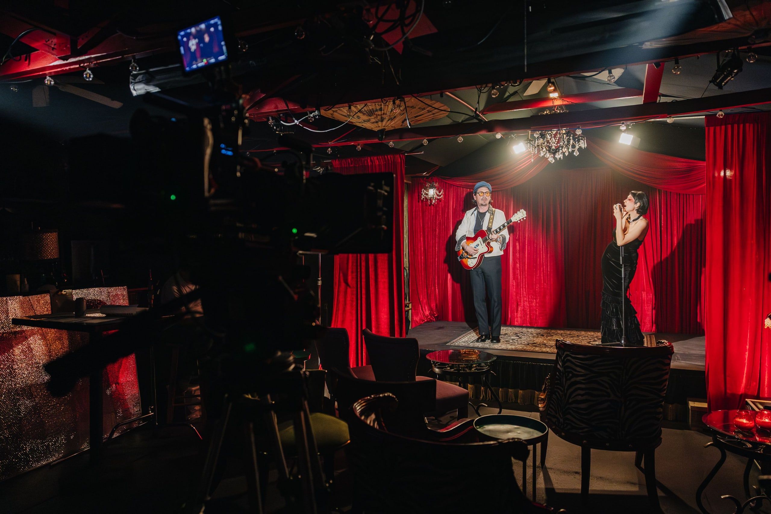 Looking past the camera at a guitarist and a singer performing on a small stage hung with red curtains