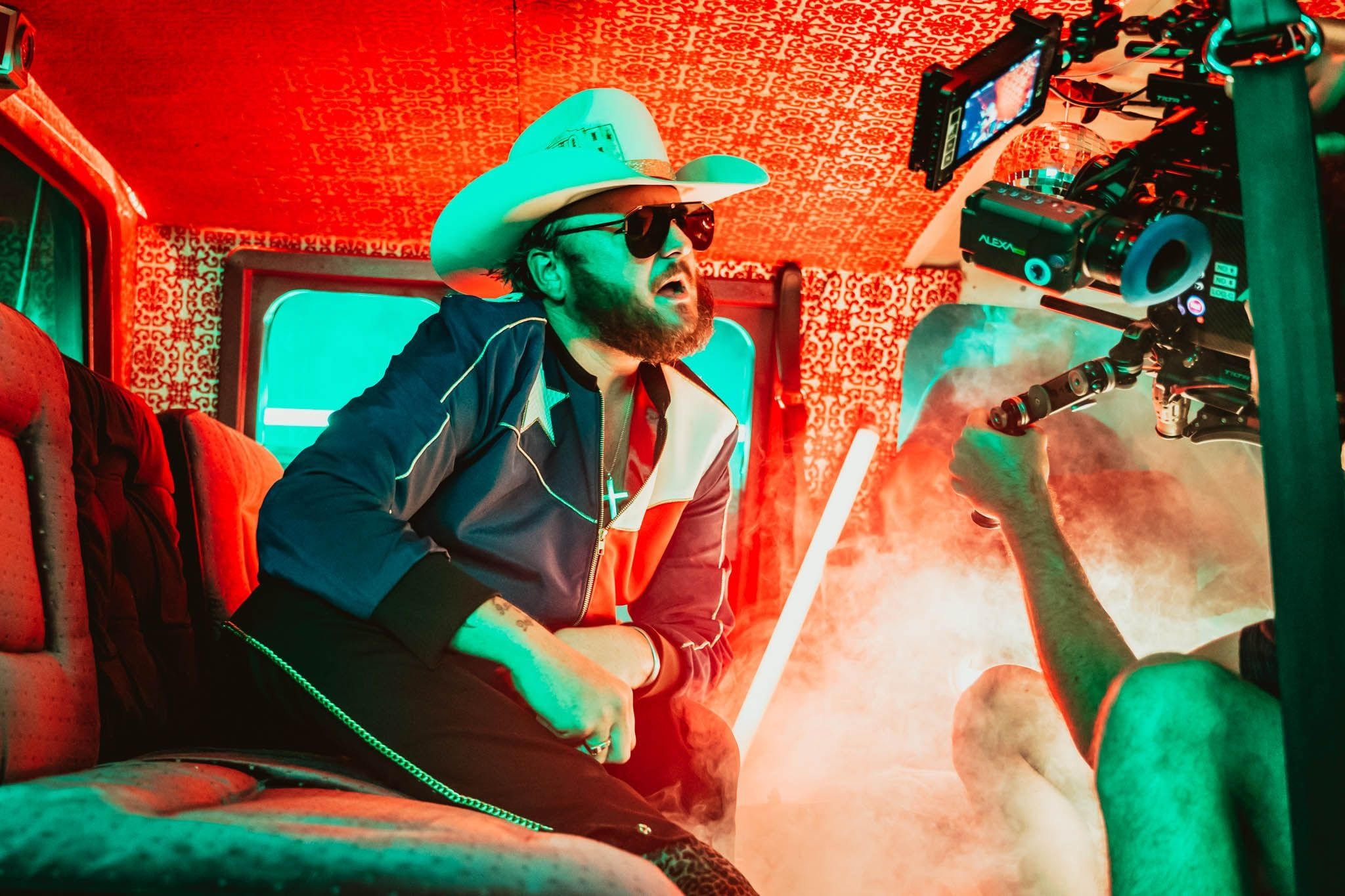 A singer in a white cowboy hat rides in a red-lit van as an operator holds the camera inches from him through the haze